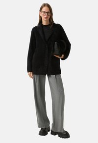 Černý huňatý pletený blazer s klopami, kombinovaný s šedými kalhotami se širokými nohavicemi. Modelka nese malou černou texturovanou tašku. Černé platformové boty.