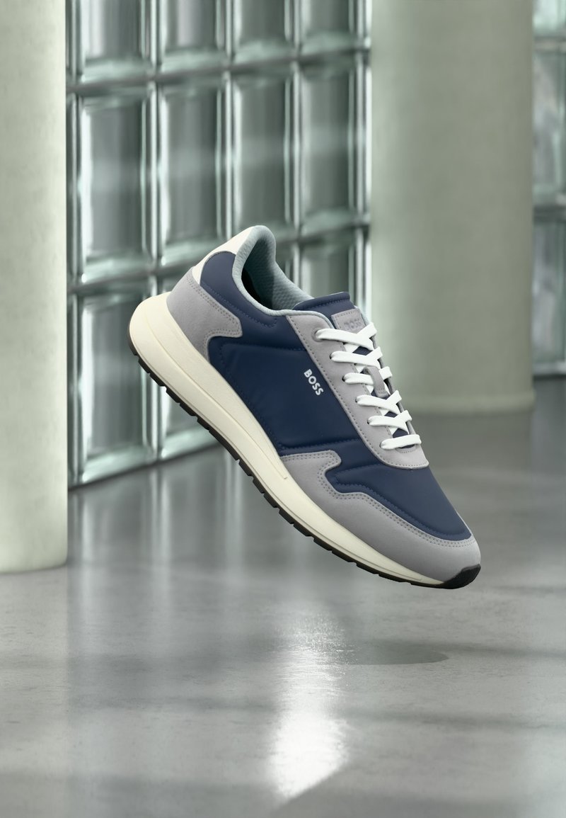 Baskets bleu marine et gris avec des lacets blancs et le logo "BOSS" sur le côté, flottant au-dessus d'un sol gris réfléchissant près d'un mur en blocs de verre.
