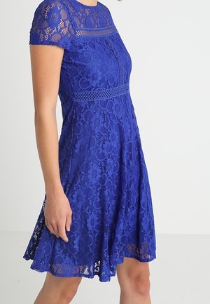 Femme portant une robe en dentelle bleu roi à manches courtes et motifs floraux, longueur genou, posant devant un fond blanc uni.