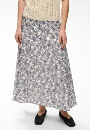Femme portant un haut en maille beige, une longue jupe crème à motif floral bleu et des chaussures noires tissées sans lacets, debout sur un fond blanc.