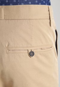 Khaki trousers with a smooth texture, featuring a back pocket secured by a dark button and a stitched seam. Partially visible is a blue patterned shirt.