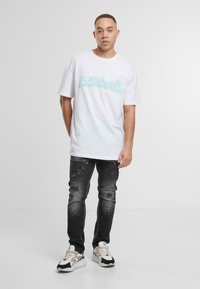 Biała bawełniana koszulka z jasnoniebieskim graficznym napisem, w połączeniu z czarnymi dżinsami o przetarciach i plamach farby. Casowe trampki.