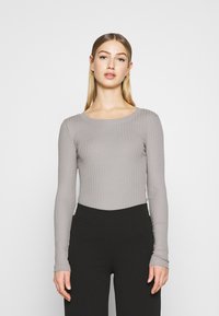 Long-sleeve, lightweight top in light gray with a ribbed texture, featuring a scoop neckline and fitted silhouette. Paired with black pants.