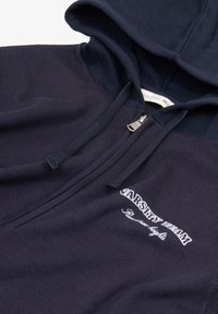 Marineblå zip-hoodie med en kontrasterende hætte, ribbet stof og "VARSITY TEAM" broderet tekst i hvid på brystet.