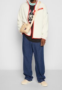 Giacca in pile bianca con dettagli rossi, maglione nero, pantaloni di jeans blu larghi, borsa a tracolla beige e scarpe da ginnastica beige. Dettagli di design semplici.