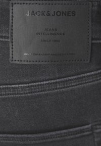 Svart läderlapp på mörk denimjeans med texten "JACK & JONES", "JEANS INTELLIGENCE", "SINCE 1990" och "CRAFTSMANSHIP AND DEDICATION."