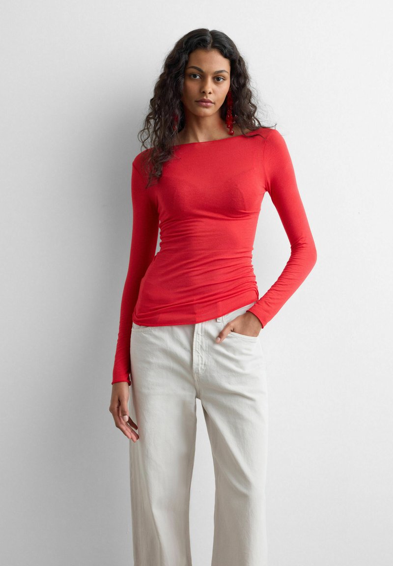 Kvinde med krøllet hår iført en tætsiddende rød langærmet top og højtaljede hvide bukser, stående mod en ensfarvet hvid baggrund.