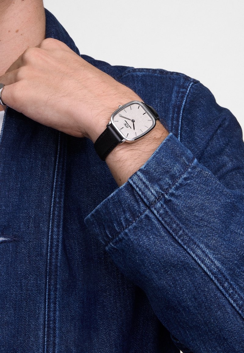 Personne portant une montre carrée en argent avec un cadran blanc et un bracelet en cuir noir, visible sur la manche d'une veste en jean bleue.