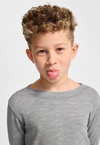 Ung person som bär en grå långärmad tröja och lekfullt visar tungan. Håret är lockigt och bakgrunden är enkel och ljusfärgad.