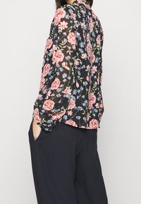 Blouse noire transparente à motif floral rose, rouge et bleu. Caractéristiques : manches longues, col, coupe décontractée, associée à un pantalon foncé.