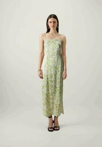 Sieviete, kas ģērbusies bezpiedurkņu, līdz potītēm garā baltā kleitā ar zaļiem lapu rakstiem, melnās augstpapēžu sandales un zelta aproce, stāv pret vienkāršu fona.