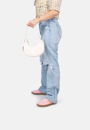 Jean déchirés bleu clair avec un ourlet effiloché, associés à des baskets roses avec des accents orange. Un petit sac à main blanc est tenu dans la main gauche.