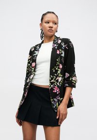Fekete virágmintás blézer rózsaszín, kék és zöld mintákkal, laza szabással és háromnegyedes ujjakkal, fehér felsővel és fekete szoknyával párosítva.