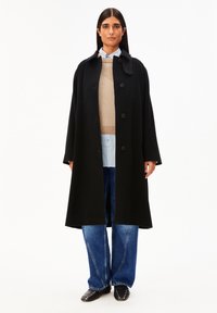 Frau trägt einen langen schwarzen Mantel über einem beigen Pullover und einem blauen Hemd, kombiniert mit blauen Jeans und schwarzen, mit Nieten verzierten Spitzen-Schuhen.