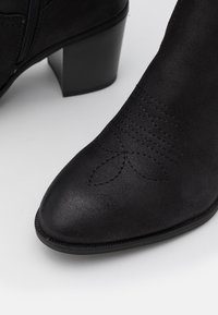 Stivale nero alla caviglia con punta a forma di rocchetto, in pelle testurizzata, e cuciture decorative sulla parte superiore. Presenta un tacco a blocco e zip laterale.
