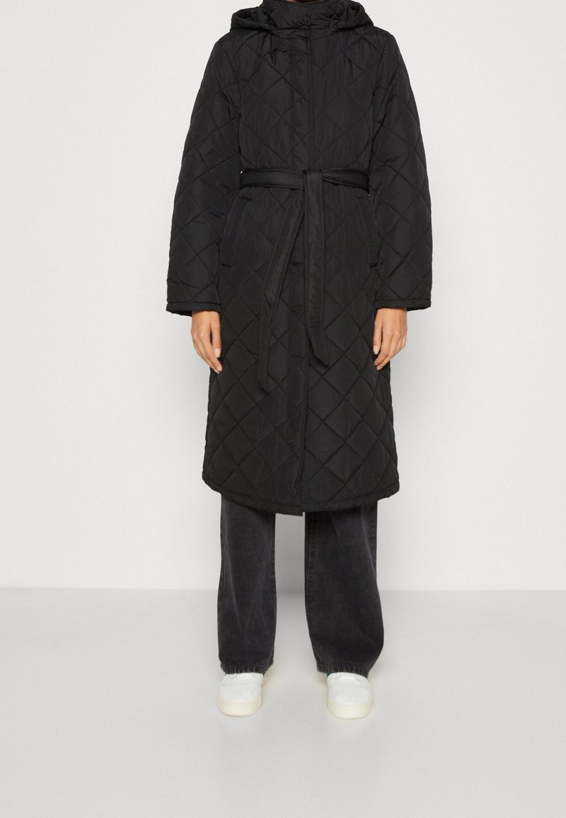 Manteau long noir matelassé avec capuche et ceinture, porté avec un pantalon noir ample et des baskets blanches, debout devant un fond clair uni.