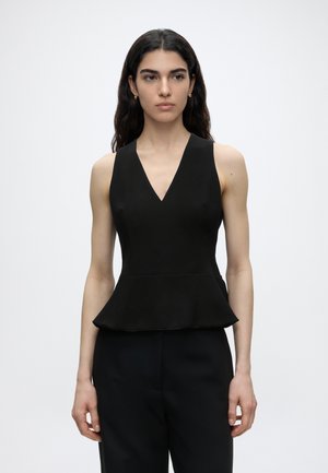 Femme aux longs cheveux foncés porte un haut peplum noir sans manches avec un col en V et un pantalon noir, debout devant un fond clair uni.