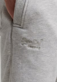 Acerca de un bolsillo de pantalones de chándal gris claro, con el logotipo bordado de SuperDry y una mano parcialmente dentro del bolsillo.