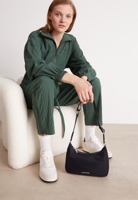 Jumpsuit verde bosco con texture, sneakers bianche con accenti beige e una piccola borsa a spalla nera con cerniera e logo.
