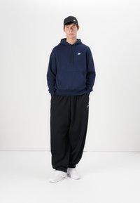 Sweatshirt azul marinho com logótipo branco, calças de fato largas pretas e ténis brancos. O boné combina com as calças, apresentando o mesmo logótipo branco.