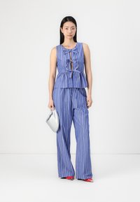 Modra in bela prugasta brezrokavna peplum bluzica z detajlom zavezovanja, v kombinaciji s matching širokimi hlačami; model drži srebrno torbico.