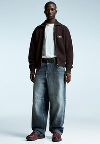 Brun stickad zip-up jacka över en vit t-shirt, tillsammans med löst sittande ljusblå denimjeans. Vinröda sneakers fulländar outfiten.