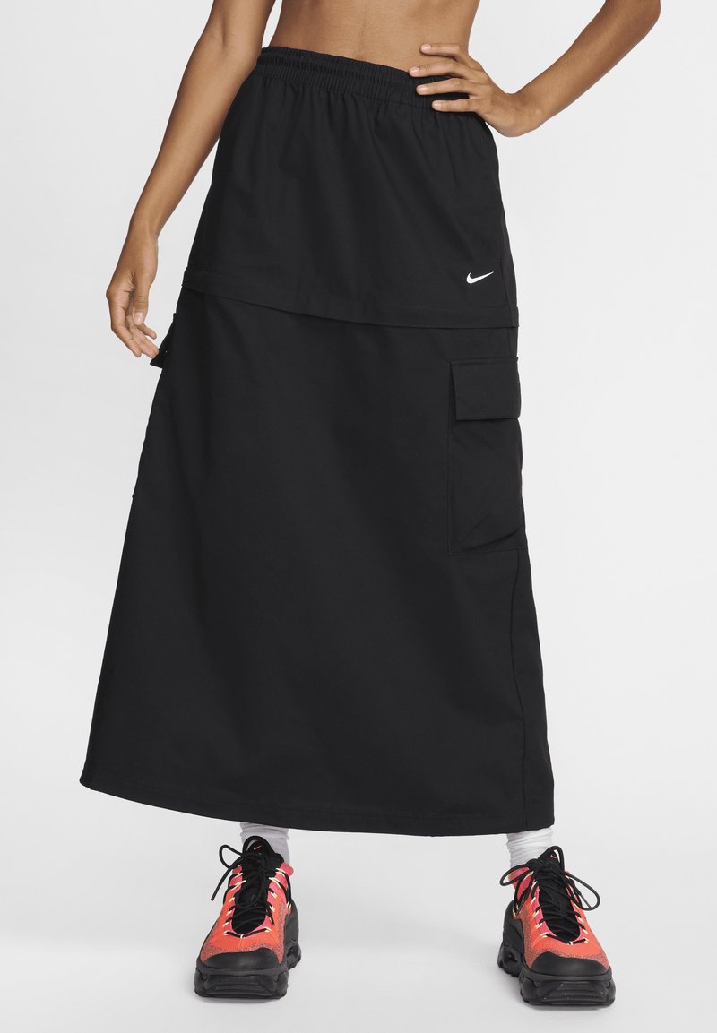Juoda sportinė ilgis sijonas su elastine juosta, su priekinėmis kišenėmis ir Nike logotipu. Dėvima su spalvingais sportiniais batais.