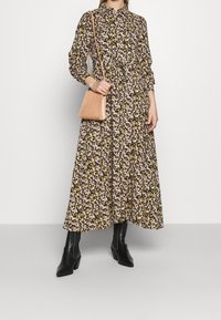 Nainen, jolla on pitkähihaisen kukkakuosisen maksimekon ylle puetut mustat polvipituiset saappaat ja joka kantaa pientä beigeä olkalaukkua.