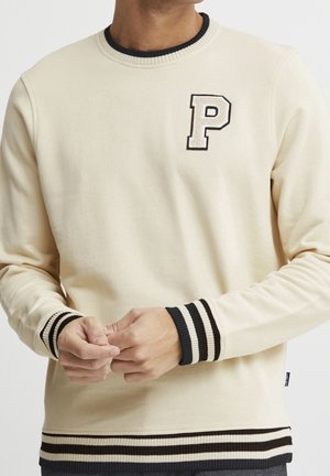 Mand iført cremefarvet sweatshirt med sorte og cremefarvede stribede ærmeafslutninger, kant og krave, med et stort bogstav P-patch på brystet.