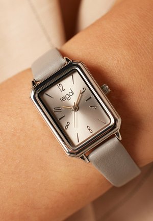 Rectangular silver watch with a grey leather strap. Features a metallic silver dial with black hour markers and brand name "regal" at the center.
