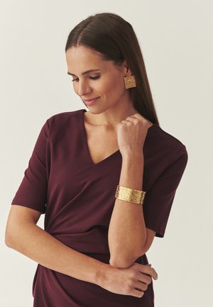 Femme en robe bordeaux portant des boucles d'oreilles carrées texturées en or et un large bracelet assorti, regardant vers le bas avec un sourire doux.