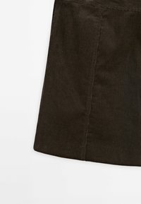 Tissu en velours côtelé marron avec des côtes verticales et une couture visible, formant une partie d'un vêtement sur fond blanc.