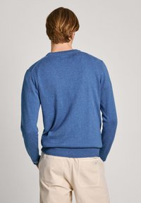 Man med kort ljusbrunt hår bär en blå långärmad tröja och beige byxor, står med händerna i fickorna vänd bort.