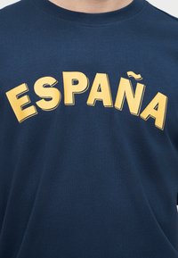Άτομο που φοράει μπλε σκούρο t-shirt με μεγάλα κίτρινα γράμματα που σχηματίζουν τη λέξη «ESPAÑA» σε καμπύλο σχέδιο στο στήθος.
