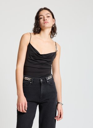 Schwarzes, figurbetontes Oberteil mit drapiertem Ausschnitt und dünnen Trägern, mit mit Perlen verzierter Hardware am linken Schulter; kombiniert mit hoch taillierten schwarzen Jeans.