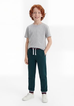 Garçon roux debout, portant un t-shirt gris clair, un jogging vert foncé avec cordon, et des baskets blanches, souriant avec une main dans la poche.