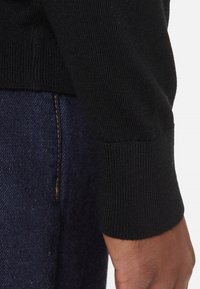 Maglione nero lavorato a maglia con polsini a costine, caratterizzato da una trama liscia. Indossato con jeans di denim scuro visibili nella parte inferiore, con una cucitura marrone.