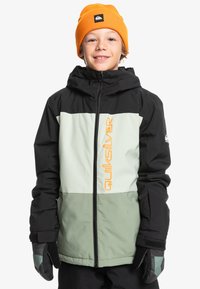 Svart och ljusgrön jacka med dragkedja fram, huva och en orange logotyp. Bärs med en orange mössa. Mjuk tygtextur.