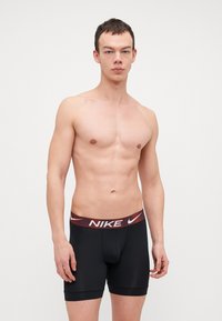 Črne Nike bokserice z rdečim in belim blagovno znamkovnim pasom, ki se tesno prilegajo in imajo gladko teksturo. Brez opaznih vzorcev ali dodatkov.