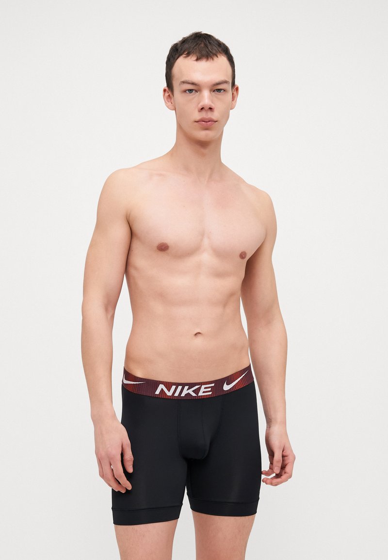 Črne Nike bokserice z rdečim in belim blagovno znamkovnim pasom, ki se tesno prilegajo in imajo gladko teksturo. Brez opaznih vzorcev ali dodatkov.
