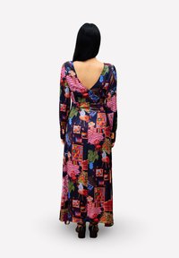 Frau mit langem dunklem Haar, die ein farbenfrohes, langärmeliges Maxikleid trägt, das Muster von Möbeln und Pflanzen zeigt, und mit dem Rücken zum Betrachter steht.