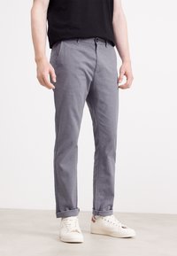 Šedé tkané kalhoty s úzkým střihem, s knoflíkem v pase a ohrnutými manžetami, doplněné bílými teniskami na neutrálním pozadí.