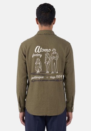 Uomo che indossa una camicia verde oliva con disegni a linee bianche di tre persone che giocano a pétanque e testo "Atomo factory, petanque cup 1954."