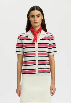 Kobieta z długimi ciemnymi włosami nosi białą, krótko rękawową koszulę zapinaną na guziki z różowo-czarnymi poziomymi paskami, czerwony szalik na szyi oraz białą spódnicę.