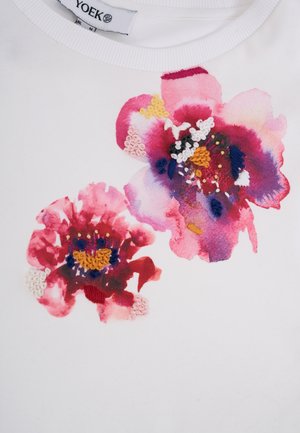 Hvid skjorte med to store pink og røde blomster i akvarelstil dekoreret med farverige broderidetaljer nær halsudskæringen.