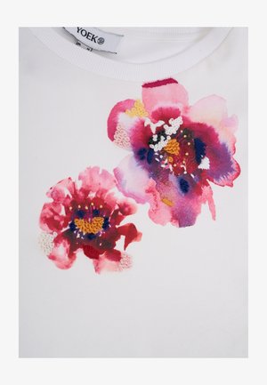 Hvid skjorte med to store pink og røde blomster i akvarelstil dekoreret med farverige broderidetaljer nær halsudskæringen.