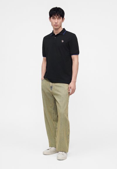 Mannequin masculin portant un polo noir avec un liseré bleu, un pantalon ample vert clair et des baskets blanches, debout avec une main dans la poche.