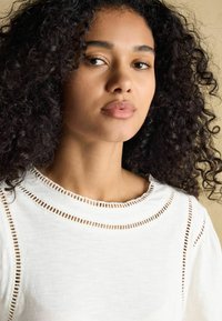 Valge kootud topp ümmarguse kaelusega, milles on avatud õmbluse detailid ääres. Pehme tekstuur, pikad varrukad ja lõdve disain.