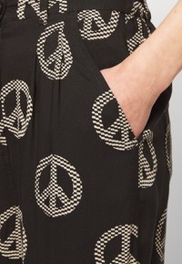 Schwarze Hose mit einem gemusterten Design großer Friedenzeichen im karierter Stil. Der Stoff wirkt glatt und leicht.