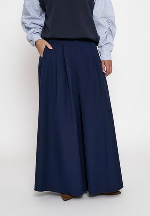 Pantaloni a pieghe ampi blu navy indossati con una camicia azzurra con motivi, una mano in tasca, mostrando scarpe marroni con punta.